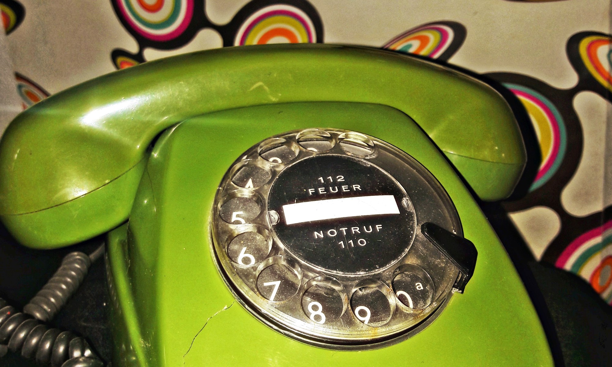 Ein grünes Wählscheibentelefon, vor dem Hintergrund einer farbenfrohen Retrotapete.