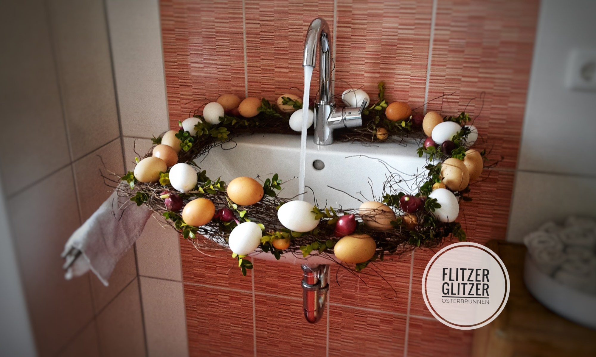 Gezeigt wird ein weißes Waschbecken mit laufendem Wasserhahn. Geschmückt ist es ringsum mit einem Kranz aus Naturmaterialien und ausgeblasenen Eiern in verschiedenen naturbelassenen Farbtönen.