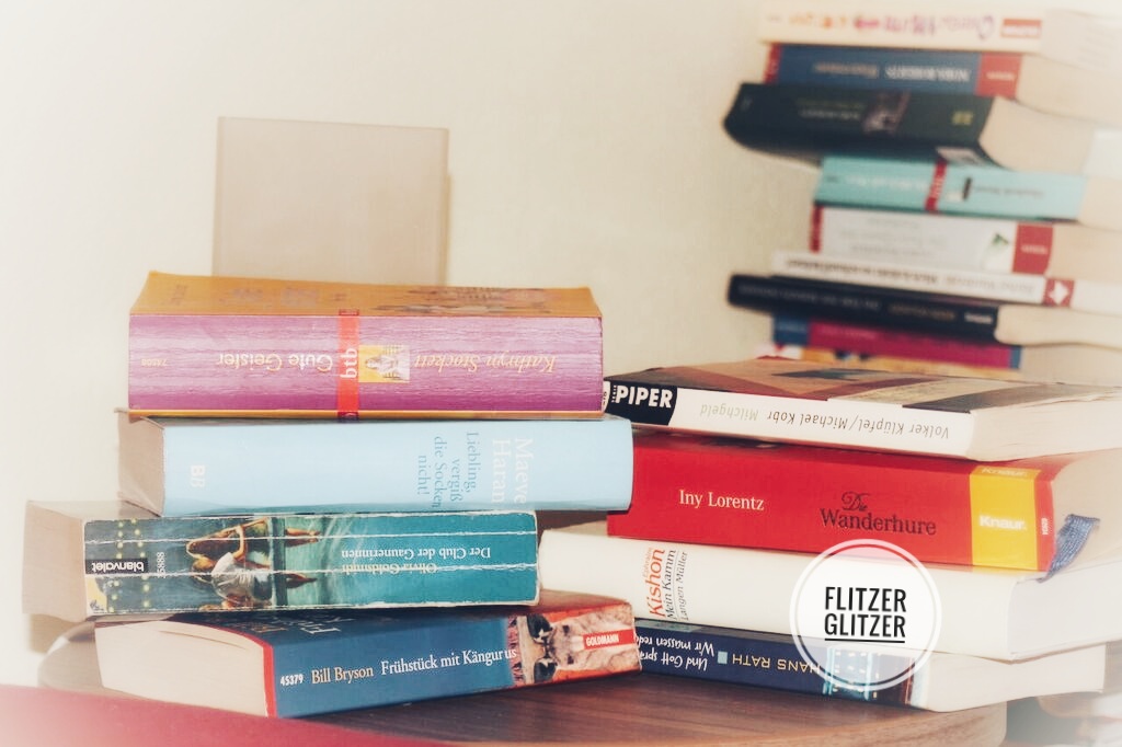 Nachttisch mit Bücherstapel. Im Hintergrund ist ein weiterer Bücherberg zu sehen.